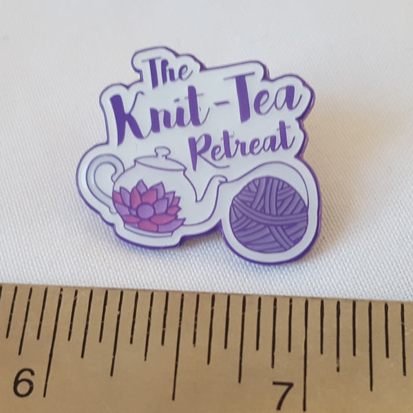Image shows a Knit-Tea Retreat pin badge featuring the logo, sitting above a metal ruler which shows that it measures approximately one inch.